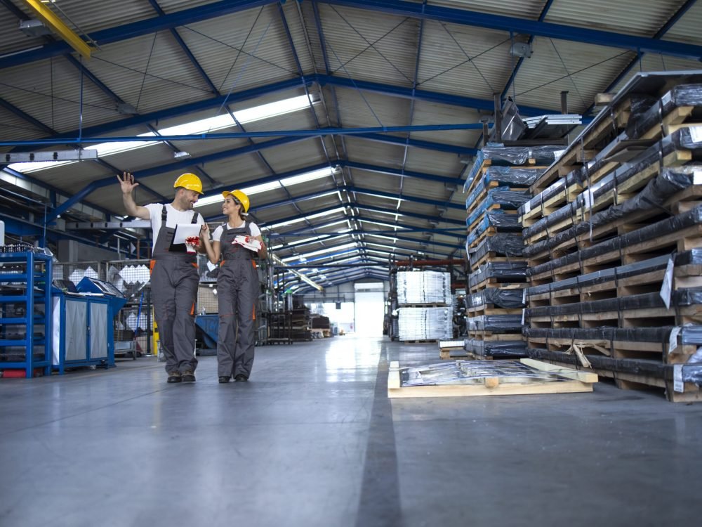 Factory workers in work wear and yellow helmets walking through industrial production hall and sharing ideas about organization.